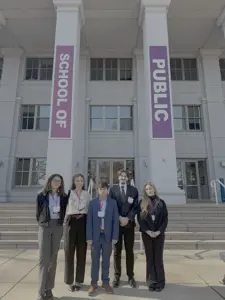A group of five students in professional dress stand in a line on the steps of a white building with three banks of windows. Two white pillars in the background have vertical banners, one reading School of and the other reading Public.