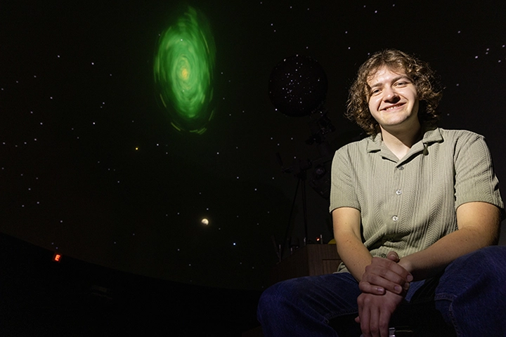 A man sits in front of a black screen that shows the night sky and a green celestial object.