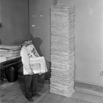 Newsboy next to giant stack of newspapers