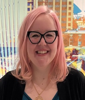 A woman with pink hair and glasses wears a black cardigan and a gold necklace.