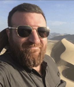 A male professor in sunglasses and a gray shirt smiles in front of sand dunes.