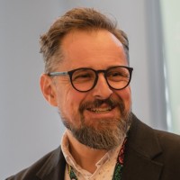 A male professor wears glasses and a suit with a white shirt and colorful lanyard.
