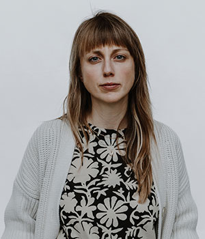 A woman with long hair and bangs wears a patterned shirt and gray cardigan.