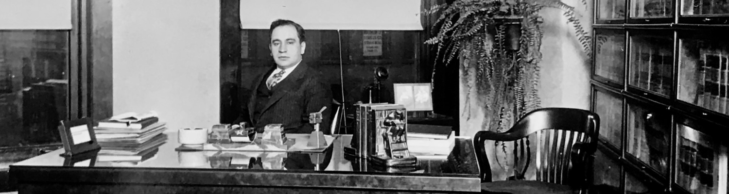 Black and white image of a man in a suit behind a stately desk.