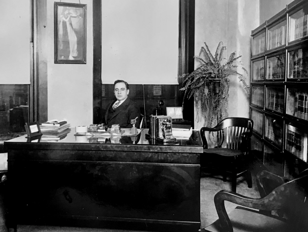Man sits behind a large desk with a window behind him and a bookcase to the side.