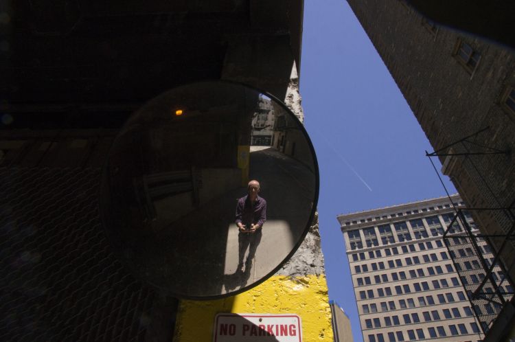 A man with a camera takes a photo of himself in a mirror in an alleyway.