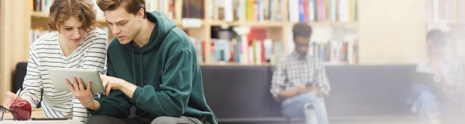 students in a library; two are looking at a tablet