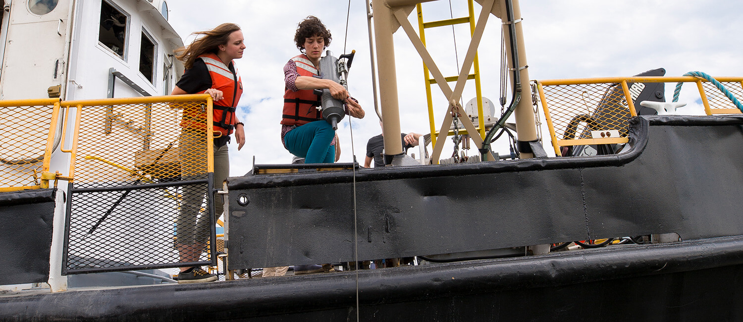 Two students (man, woman, both white) lowering a buoy into the water from the Neeskay