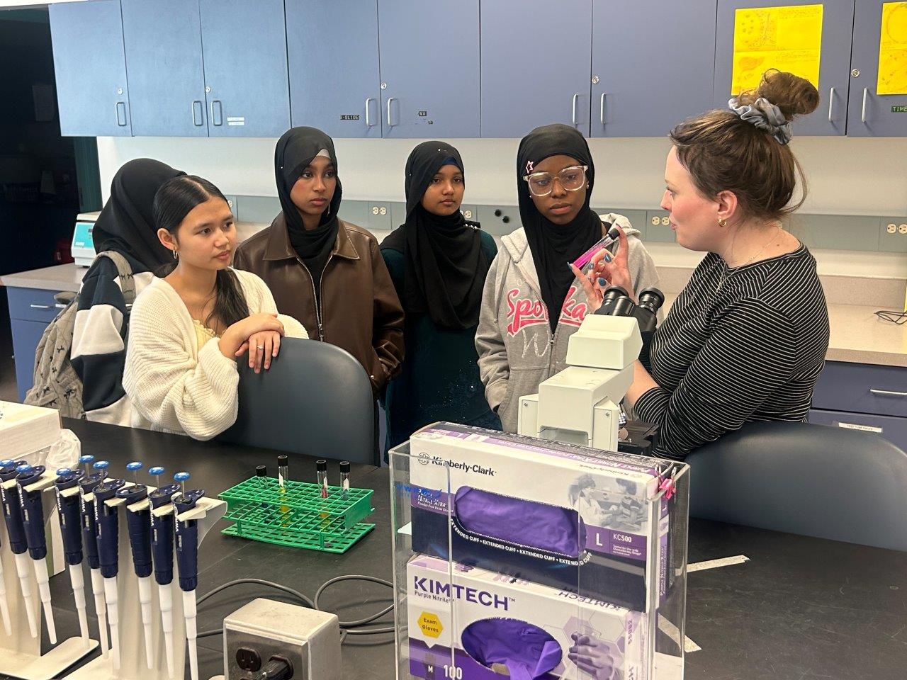 Five students gathered around a science lab table, talking about using a compound light microscope.