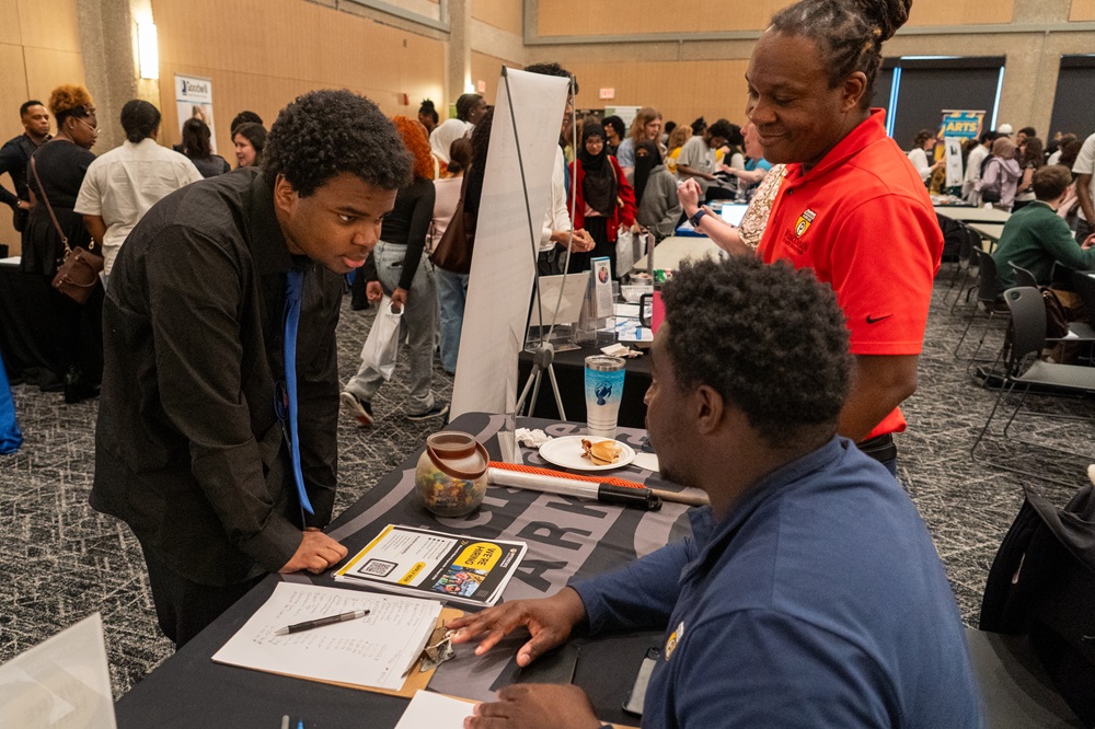 Three people stand at a table during a crowded indoor student job fair. One person leans forward to review a clipboard while two others behind the table gesture toward printed materials, with display signs, flyers, and other attendees visible throughout the room.