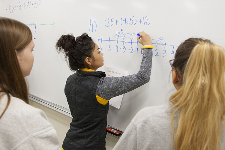 a teaching assistant demonstrating answers to a solution on the white board in front of two students 