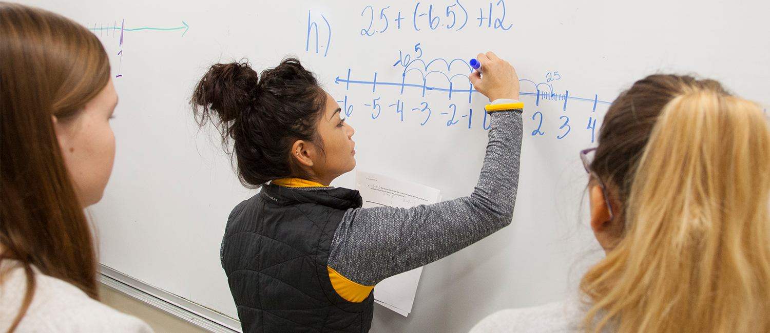 A student writes a math problem on a white board.