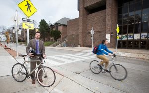 Professor stands next to his bike while another bicyclist whizzes past.
