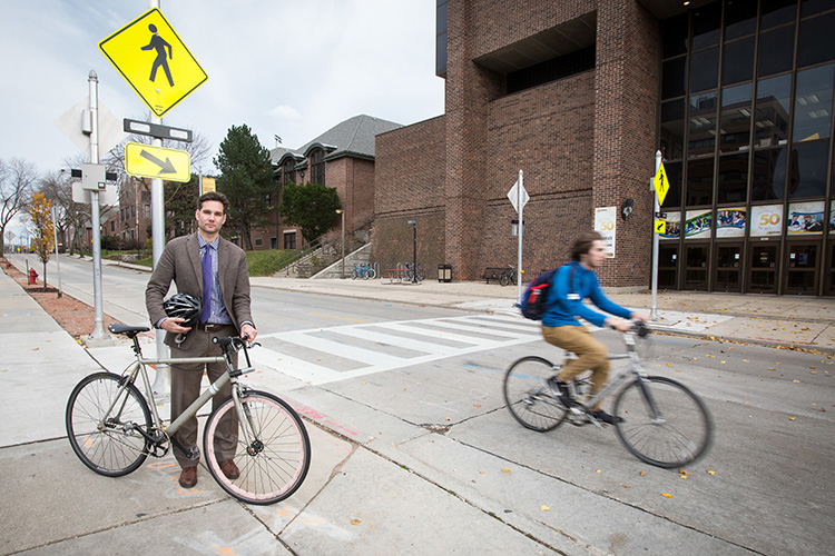 Professor stands next to his bike while another bicyclist whizzes past.