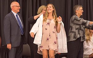 A nursing student puts on a white coat with help from another woman.