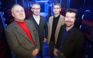 Four men stand in a room full of computers.