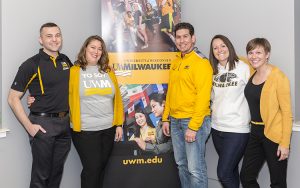 Five people stand and pose for a photo in front of a UWM poster.