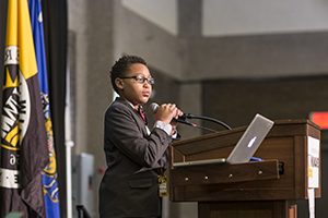 A boy stands at a podium on a stage addressing a large audience. 
