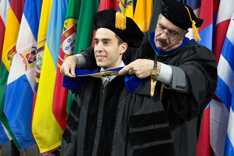 Mohsen, wearing regalia, places a hood over a student in his graduation cap and gown