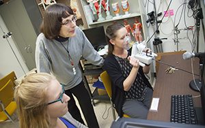 Two students sit at computers while a teacher stands behind them.