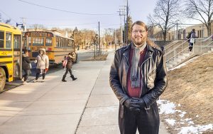 Marcus Britton stands on a sidewalk in front of a school as children disembark from the buses behind him.