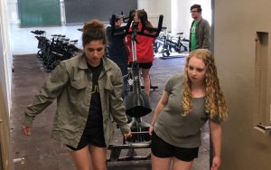 Two girls carry an exercise bike out of a brightly lit room