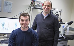 A professor stands behind a seated student in a lab.