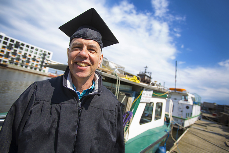 A man stands in a graduation gown and cap in front of a boat.
