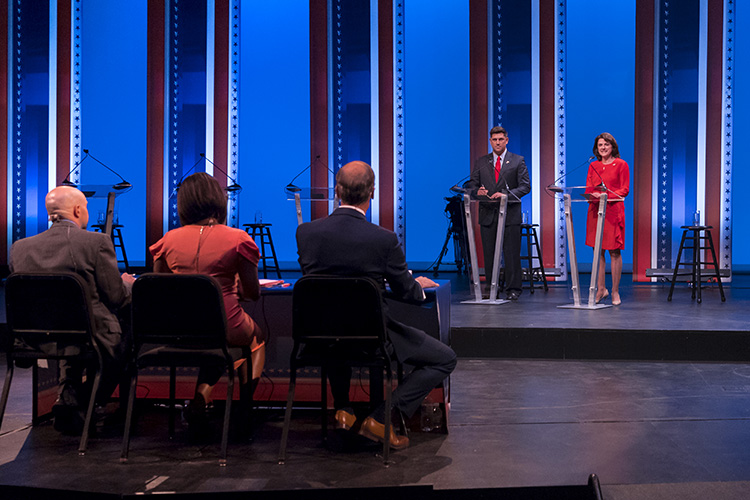 The two candidates stand onstage in front of podiums, while three debate moderators sit at a table with their backs tot he camera.