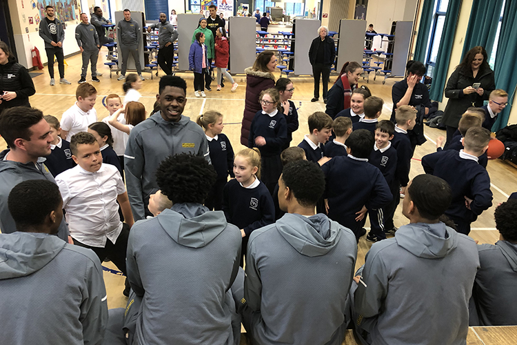 Basketball players sit on stage with their backs to the camera while elementary students sit on the floor and ask questions.