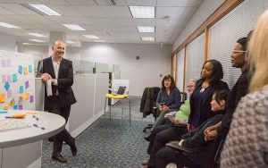 A man stands at a whiteboard while others sit and watch him.