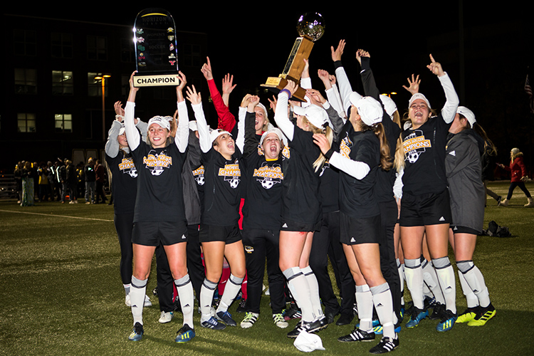 Players stand on the soccer field holding aloft two championship trophies.