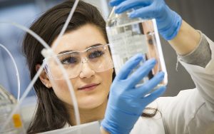 A woman wearing gloves and safety glasses lifts a beaker and looks at it.