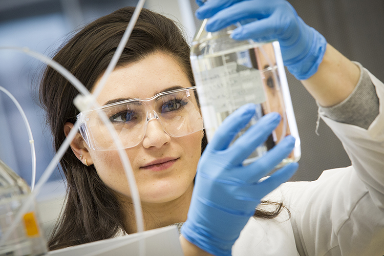 A woman wearing gloves and safety glasses lifts a beaker and looks at it.