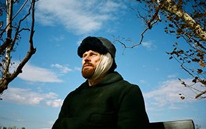 Willem Dafoe stares off to the left with a blue sky behind him.