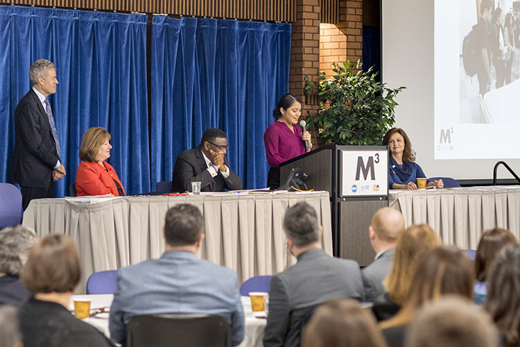 A woman speaks at a podium while others sit at a table flanking her.