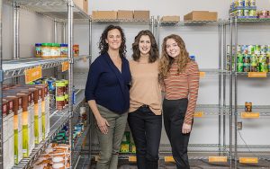Three women stand among shelves of food.