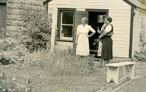 A black and white photo shows three women standing near a garden near a shed.