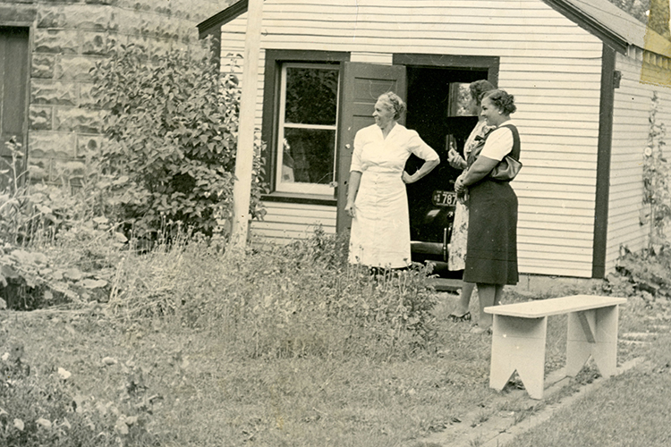 A black and white photo shows three women standing near a garden near a shed.