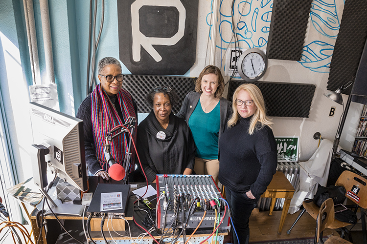 Four women pose for a photo behind a radio station desk.