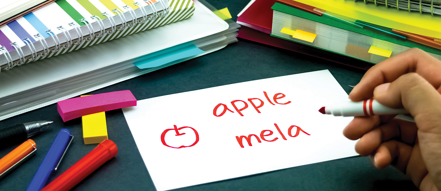 Books and other school supplies, with a hand writing 'apple mela' on a notecard