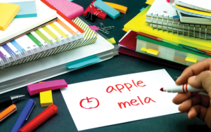 Books and other school supplies, with a hand writing 'apple mela' on a notecard