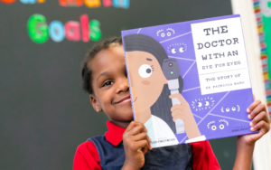 A young girl smiles and holds up a book, covering half of her face