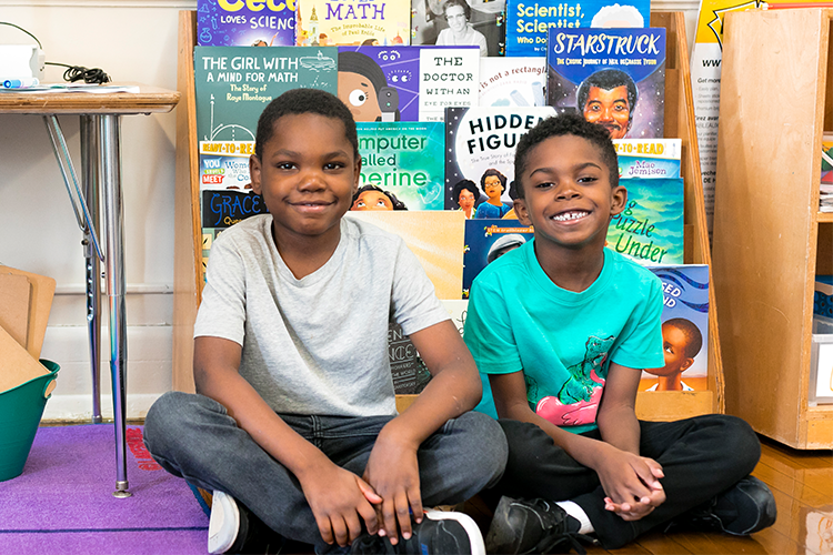 Two young boys smile and sit on a floor in front of a bookcase