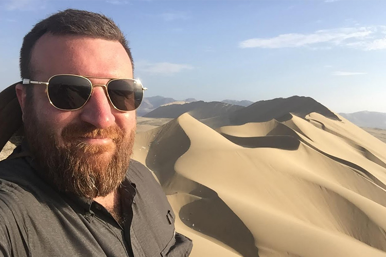Man wearing sunglasses poses in front of a sand dune.