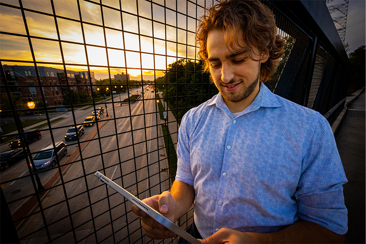 A man stands on a pedestrian bridge over a city street busy with traffic. He's holding and looking at an ipad.