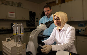 Rashida Parveen in a lab looking through a microscope with her faculty mentor nearby