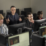 The image shows three students watching video on computer screens as UWM Associate Professor Aragorn Quinn provides coaching. Quinn's hands are raised and you can see him speaking.