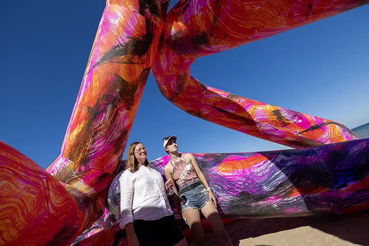 Two women stand on a beach as a colorful, tubelike inflatable art sculpture towers over them.