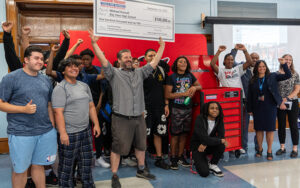 A man in a gray shirt holds an oversized check for $100,000 above his head, surrounded by several high school students and adults.